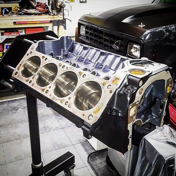 Engine block mounted on a stand in a garage, painted black with silver cylinders, next to a black truck.