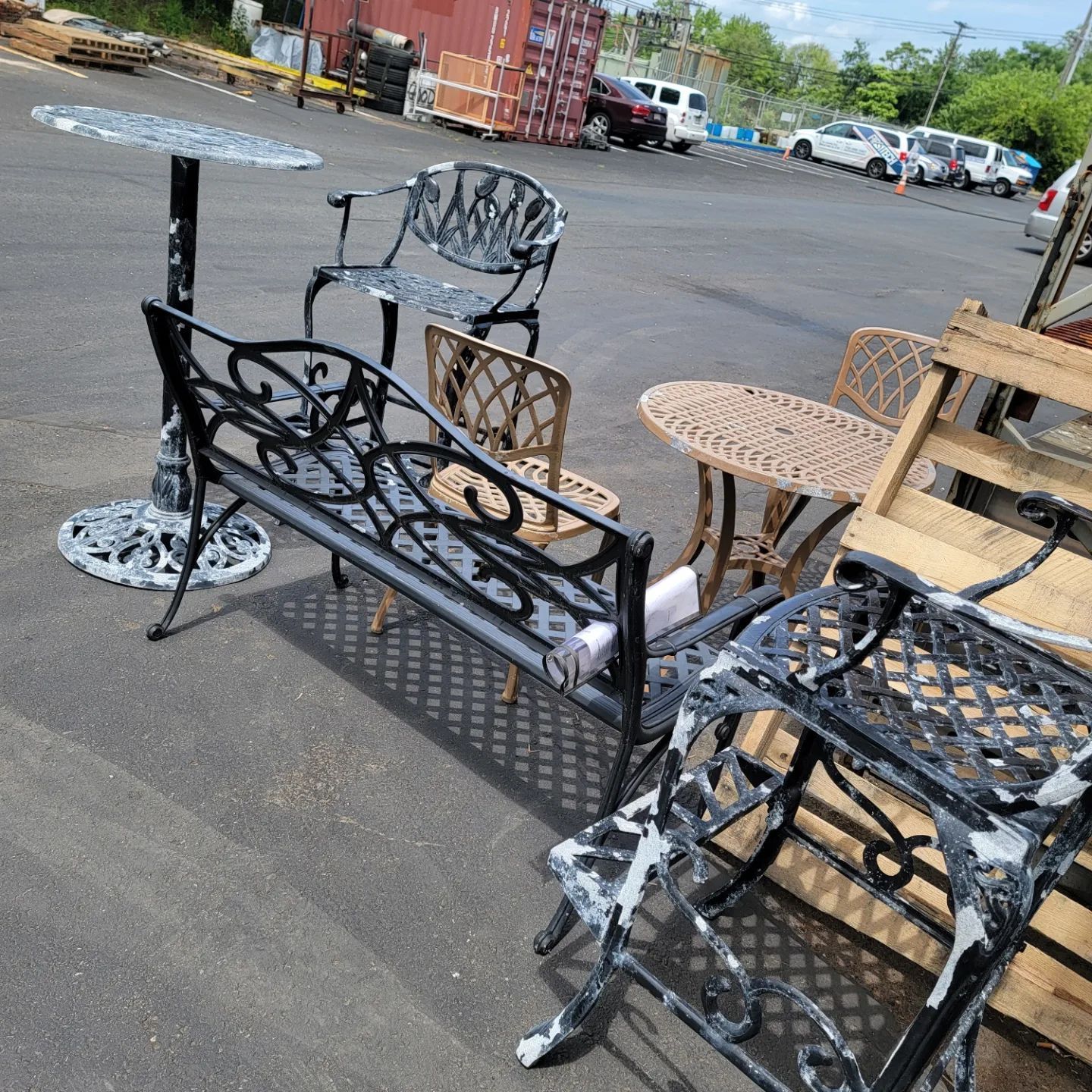 Outdoor furniture set on asphalt in a parking lot. Includes tables, chairs, and a bench, some with peeling paint.