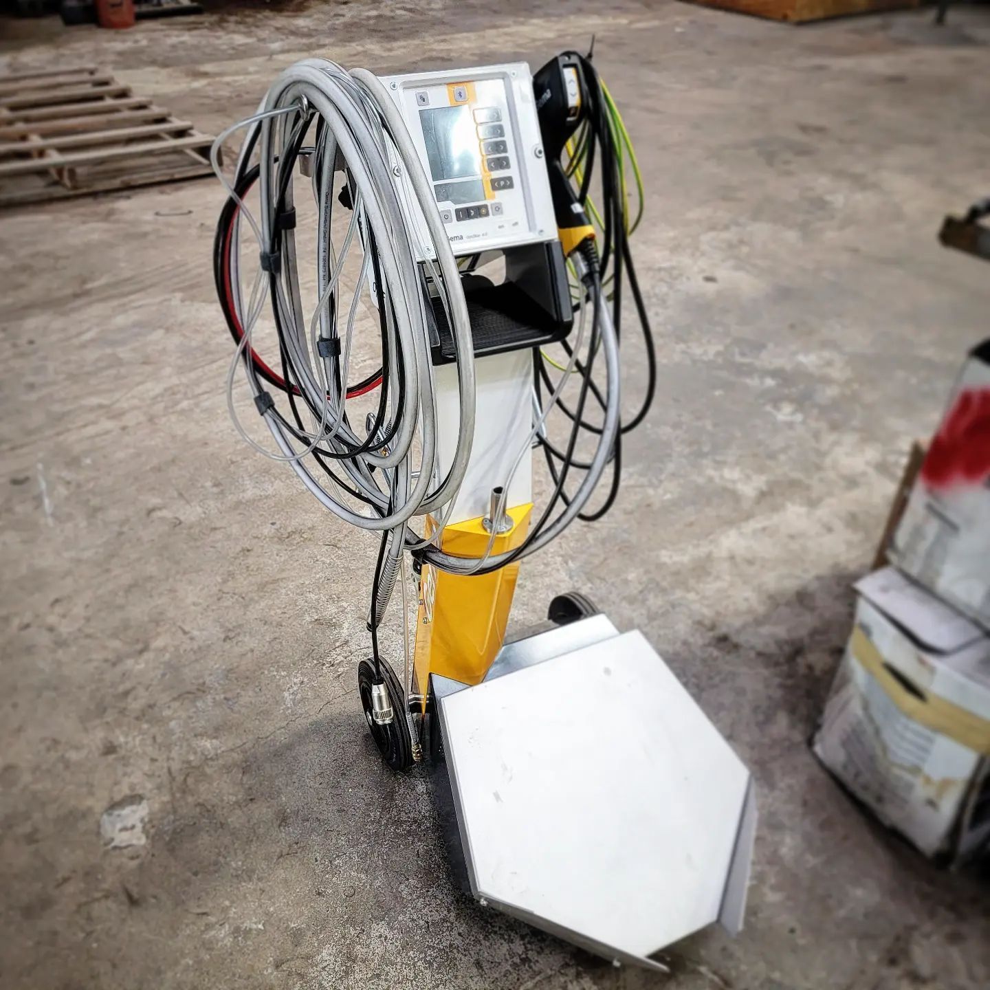 Powder coating machine with cables, controls, and yellow and white paint on a concrete floor.