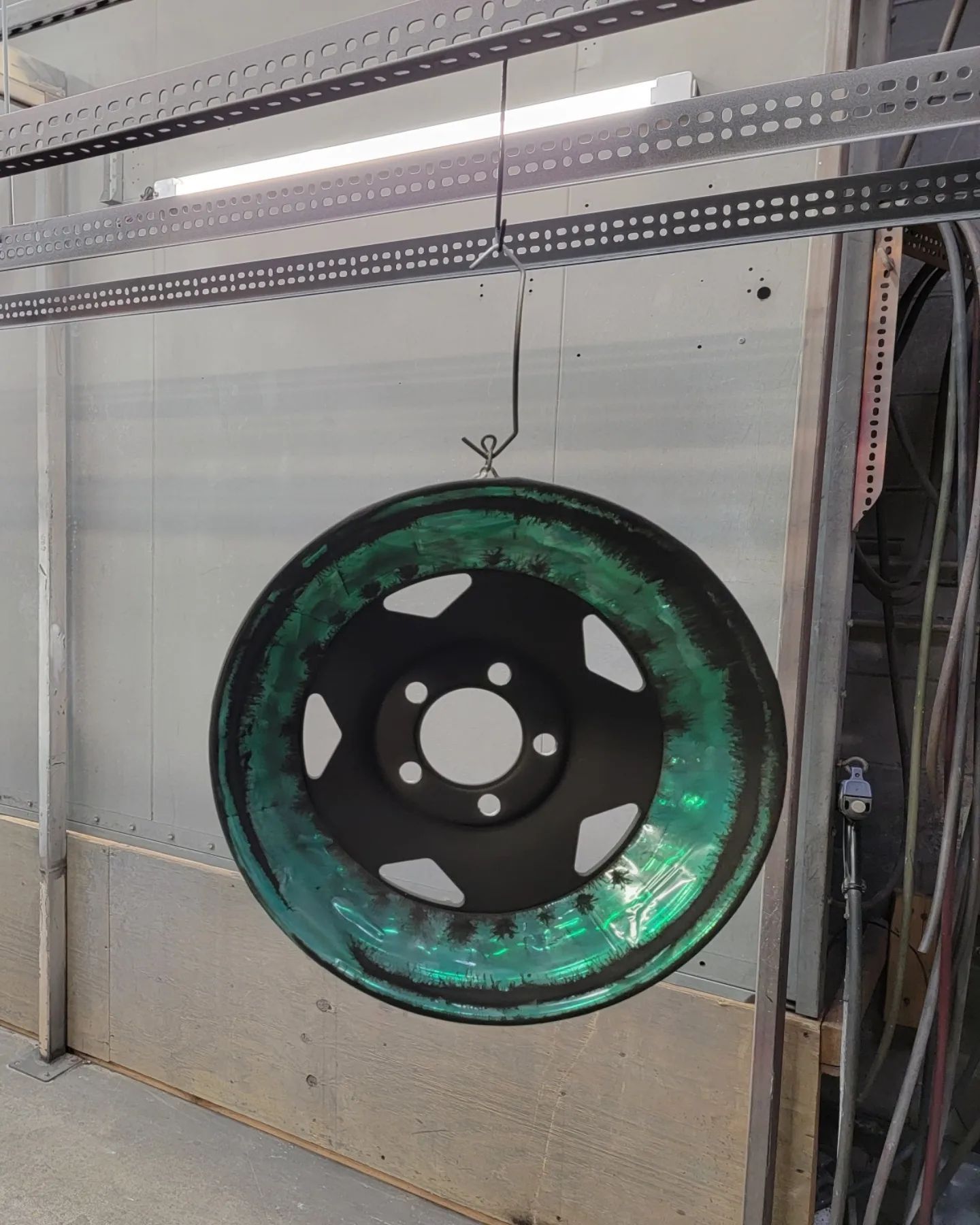 A black car wheel with iridescent teal coating hanging in a spray booth.