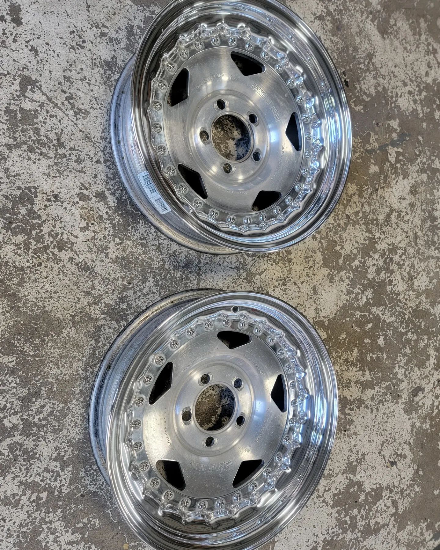 Two polished silver car wheels on a textured gray surface. The wheels have a star-shaped center and a decorative outer edge.