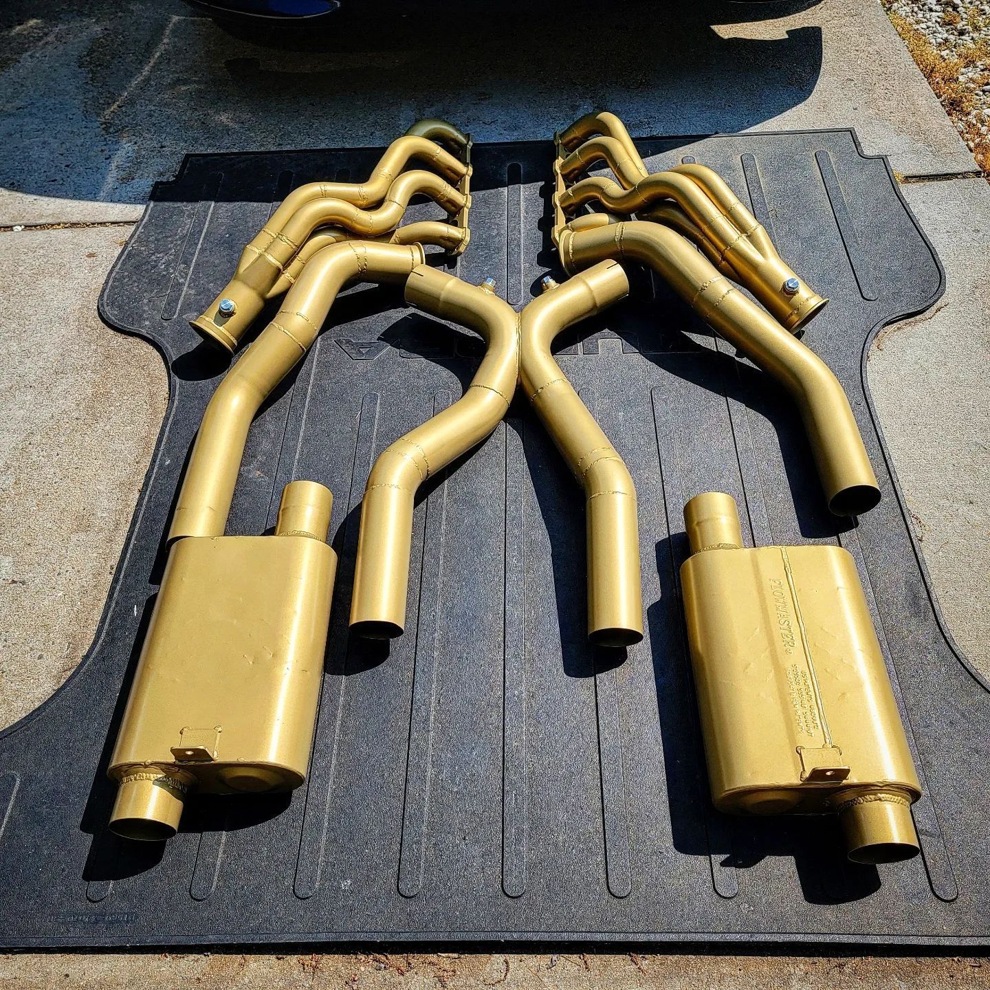 Golden car exhaust system laid out on a black mat outdoors.