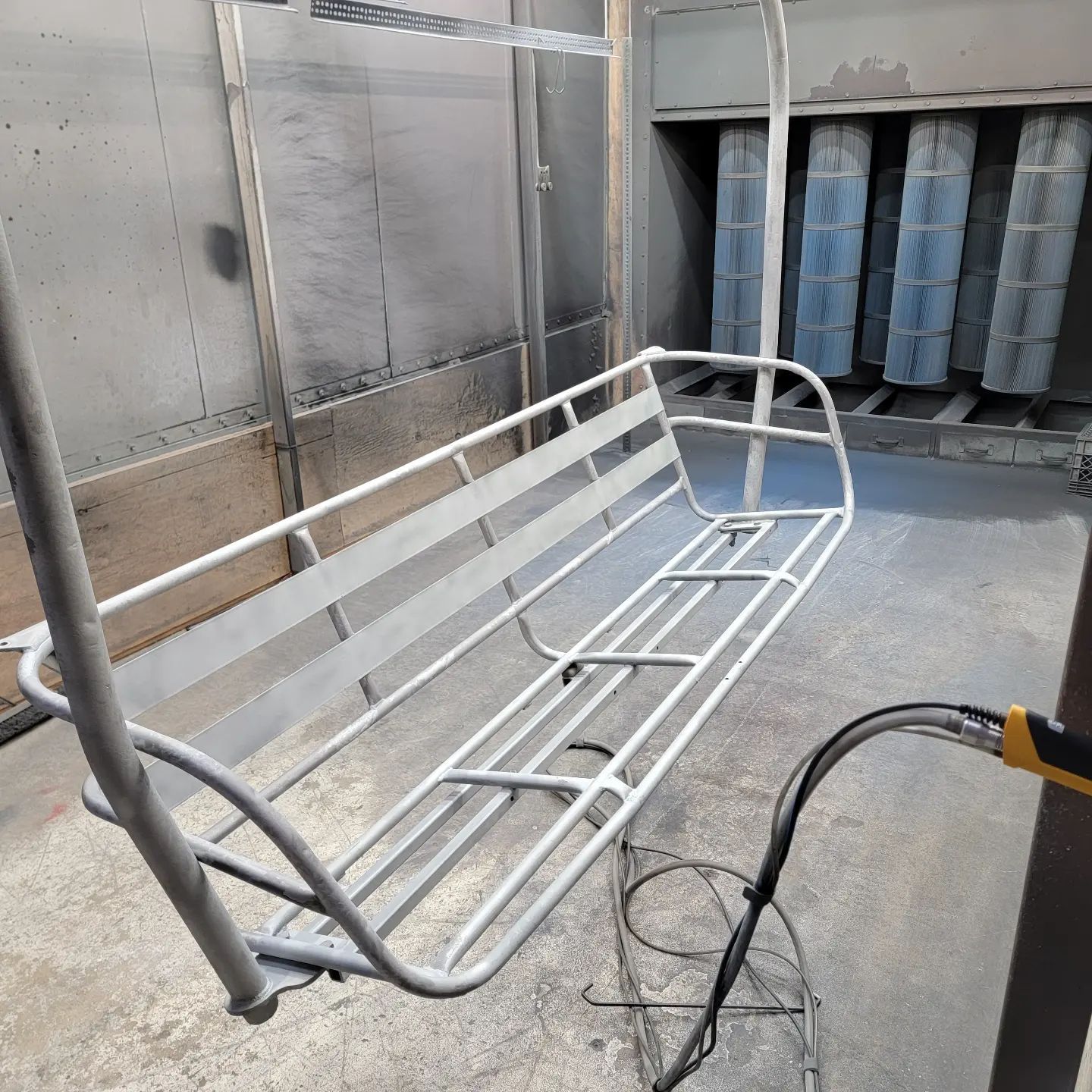 Metal bench frame inside a spray booth. The bench is being painted gray.