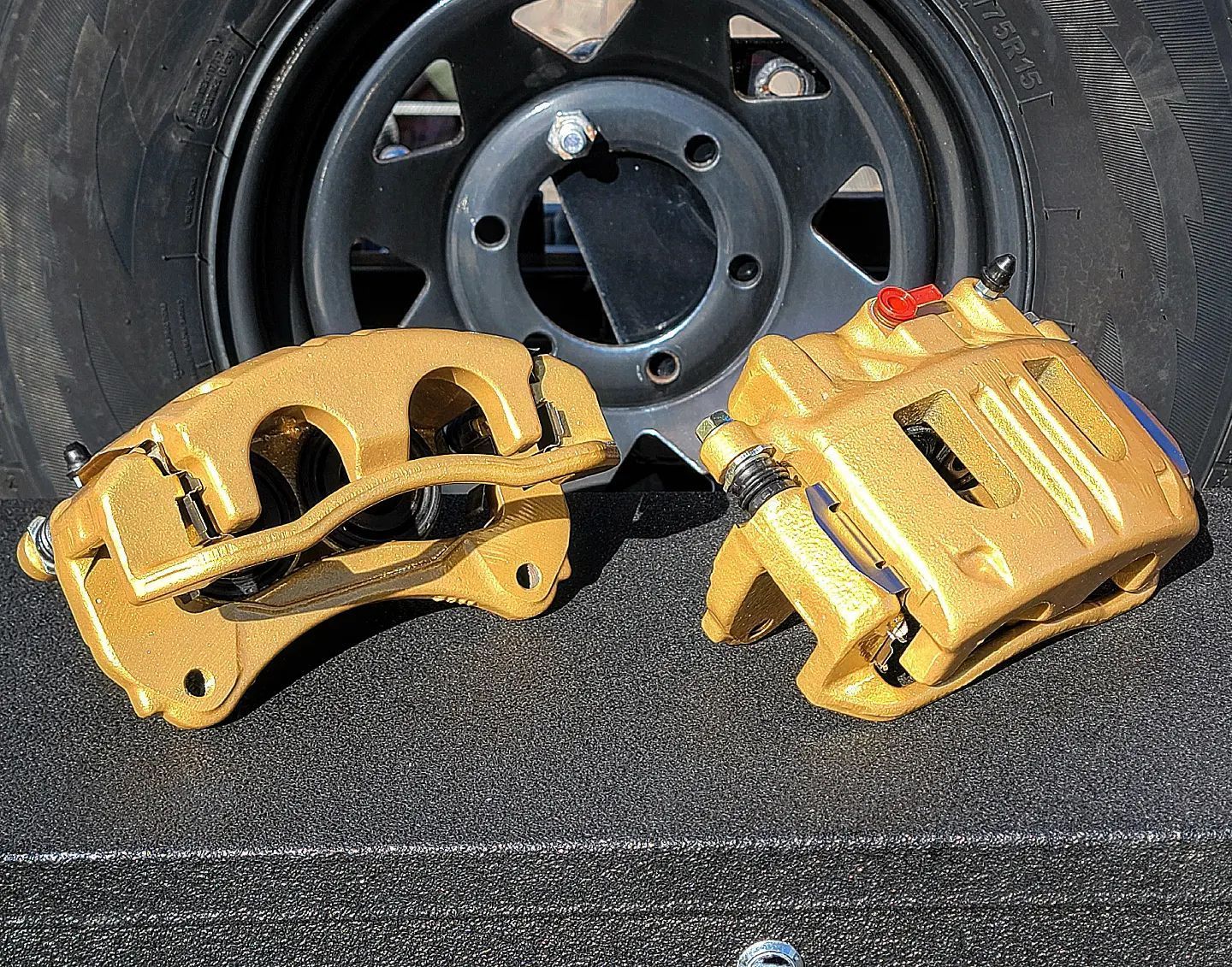 Two gold-colored brake calipers in front of a black wheel and tire, sitting on a textured surface.