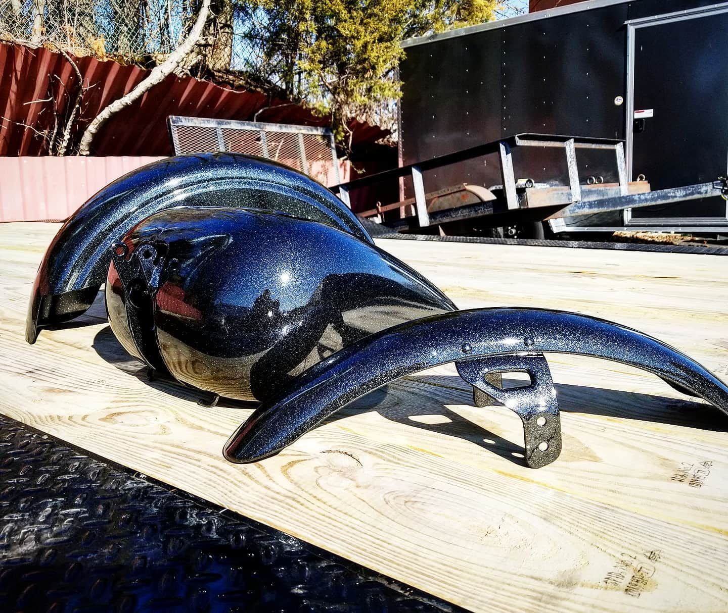Black motorcycle fenders and tank on a wooden surface, with a black trailer in the background.