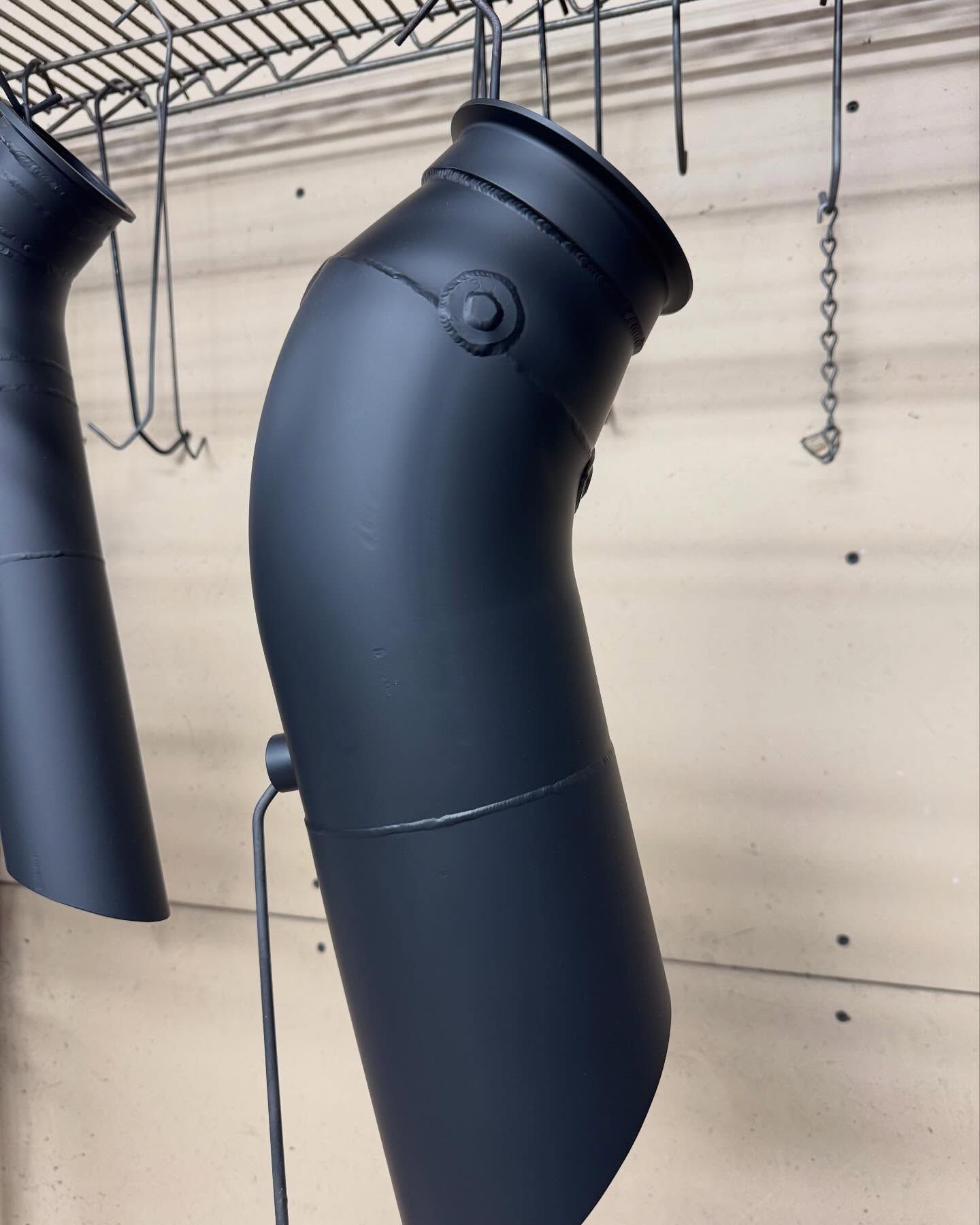 Black painted metal exhaust pipe hanging in a workshop, attached to a wire rack.