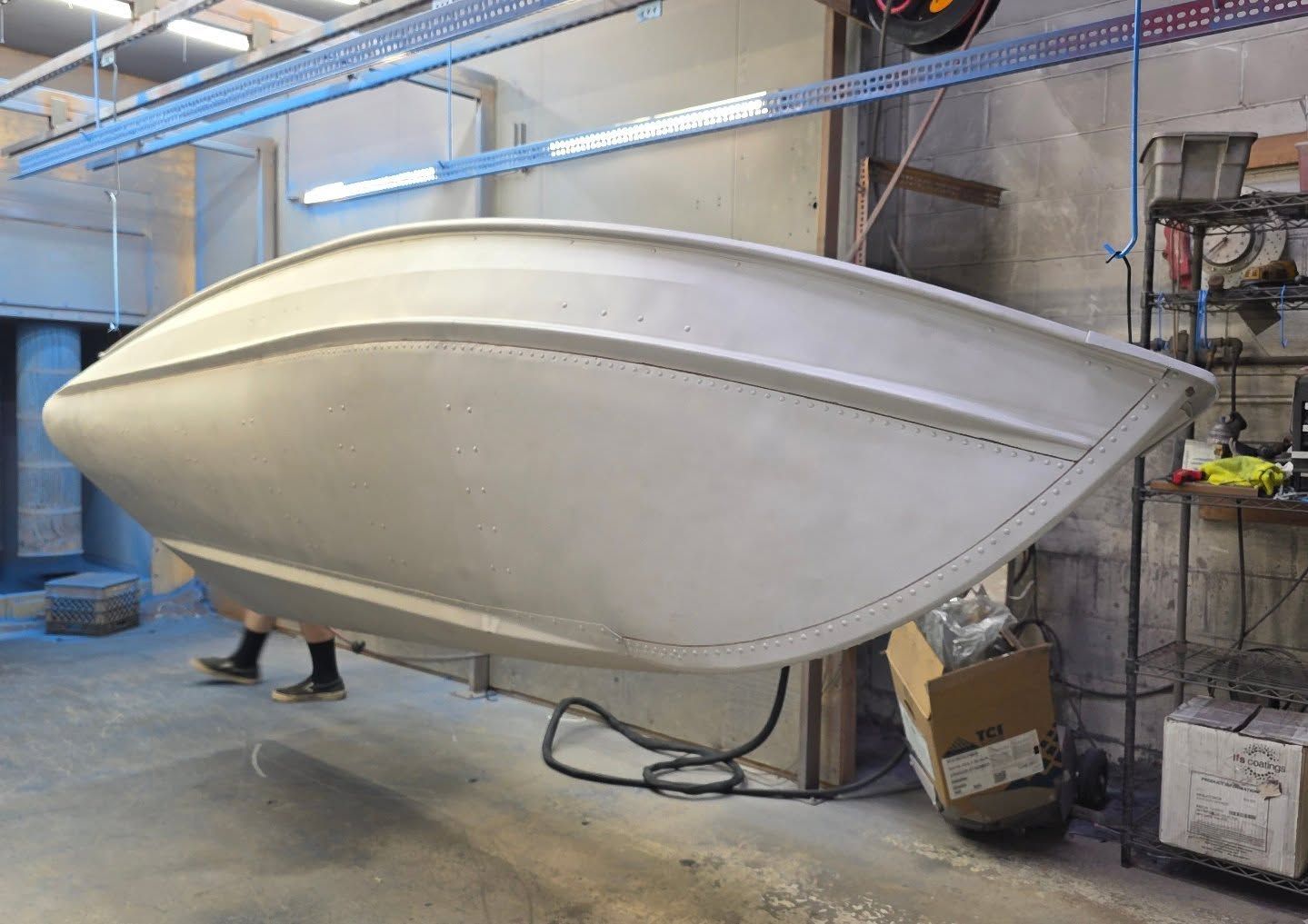 Boat hull, upside down, in a workshop. Light gray, suspended by cables, being worked on.
