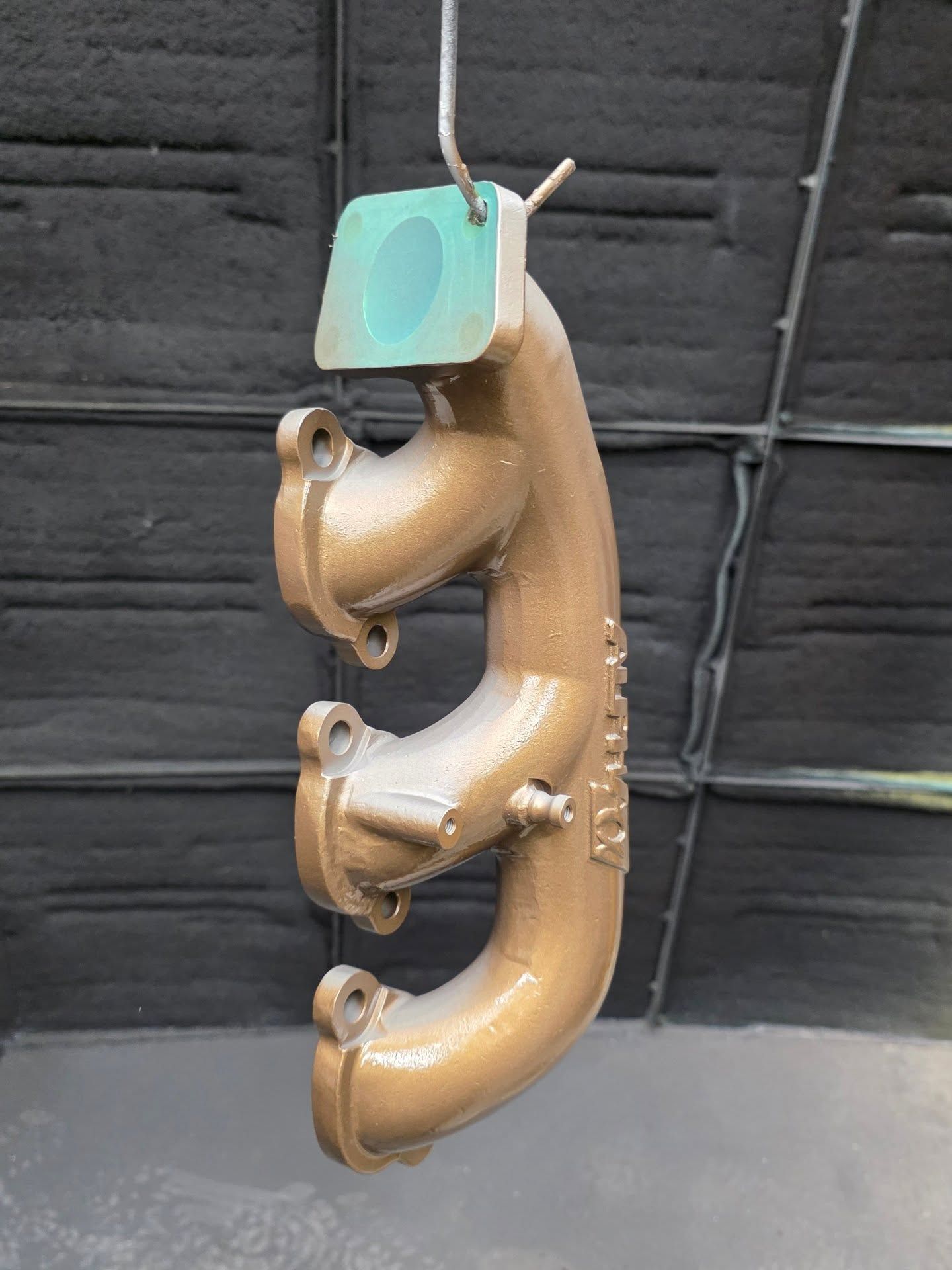 Brown painted metal exhaust manifold, hanging from a wire in a spray booth.