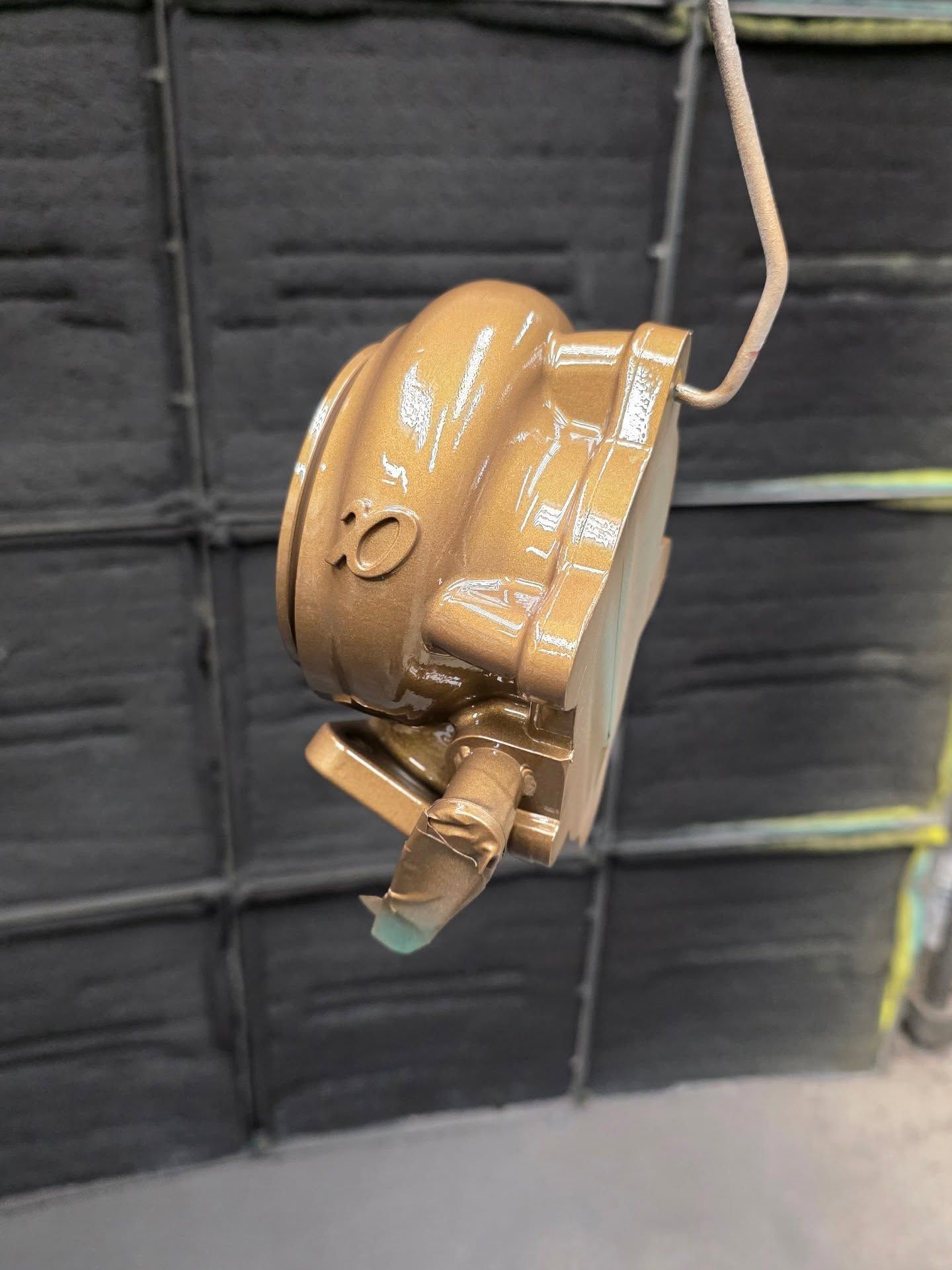 Gold-painted metal automotive part hanging from a wire in a paint booth.