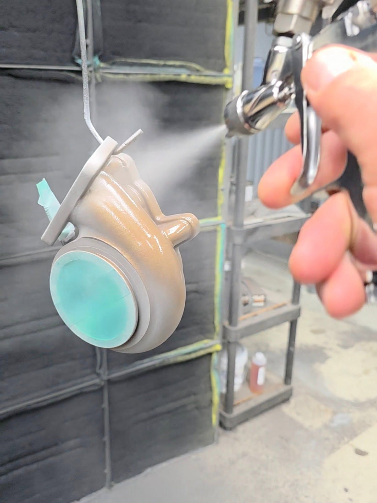 A person sprays a beige, teal, and silver-colored object with a spray gun inside a workshop.