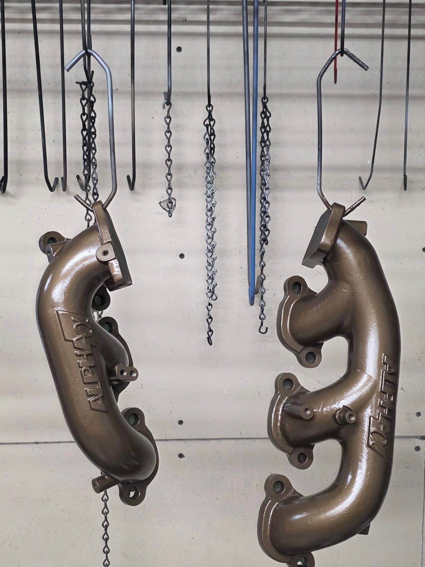 Two bronze-colored exhaust manifolds hang from chains, likely freshly coated, against a neutral backdrop.