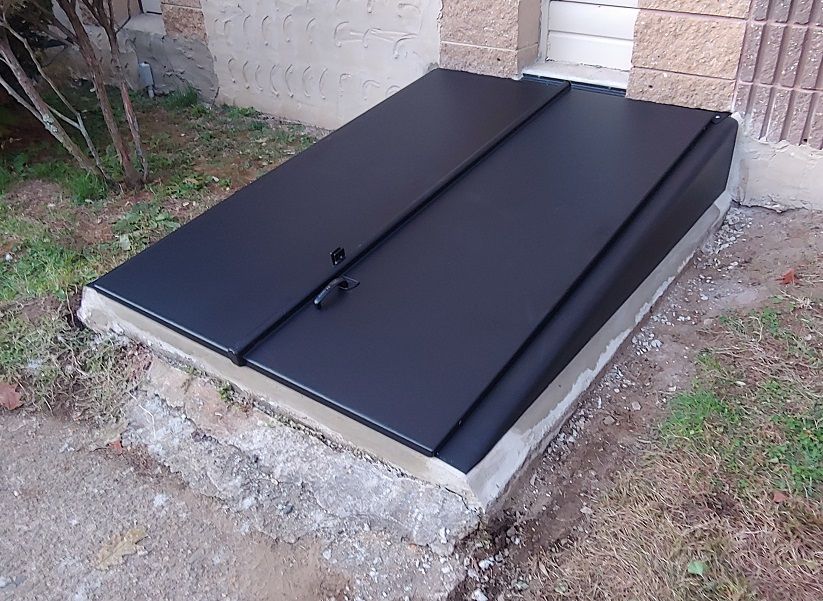 Black metal cellar doors on a concrete base, set into the ground near a building.