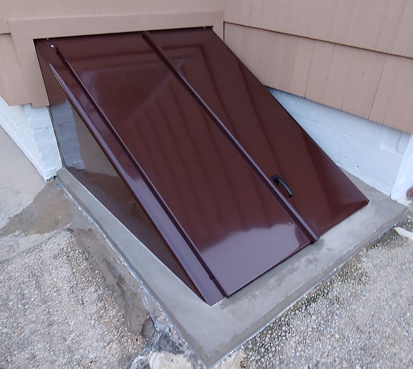 Brown metal cellar door built into concrete at the base of a tan building.