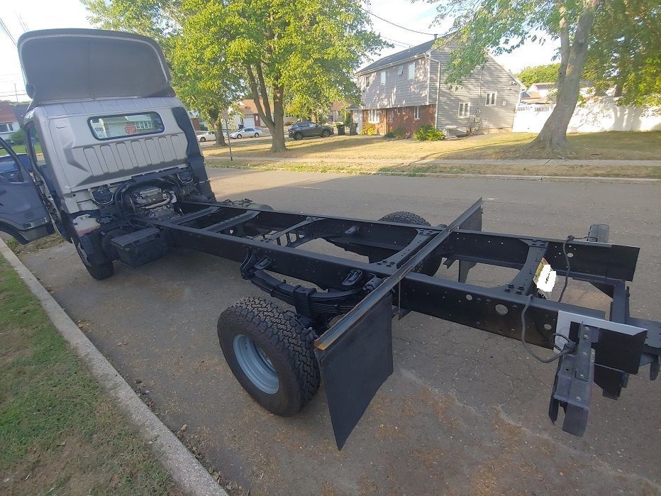 Gray truck chassis on a paved roadside. The truck bed is removed. Trees and houses are in the background.