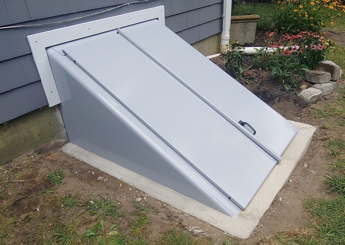 Gray sloped basement door with handle, set in concrete, next to a house with gray siding.