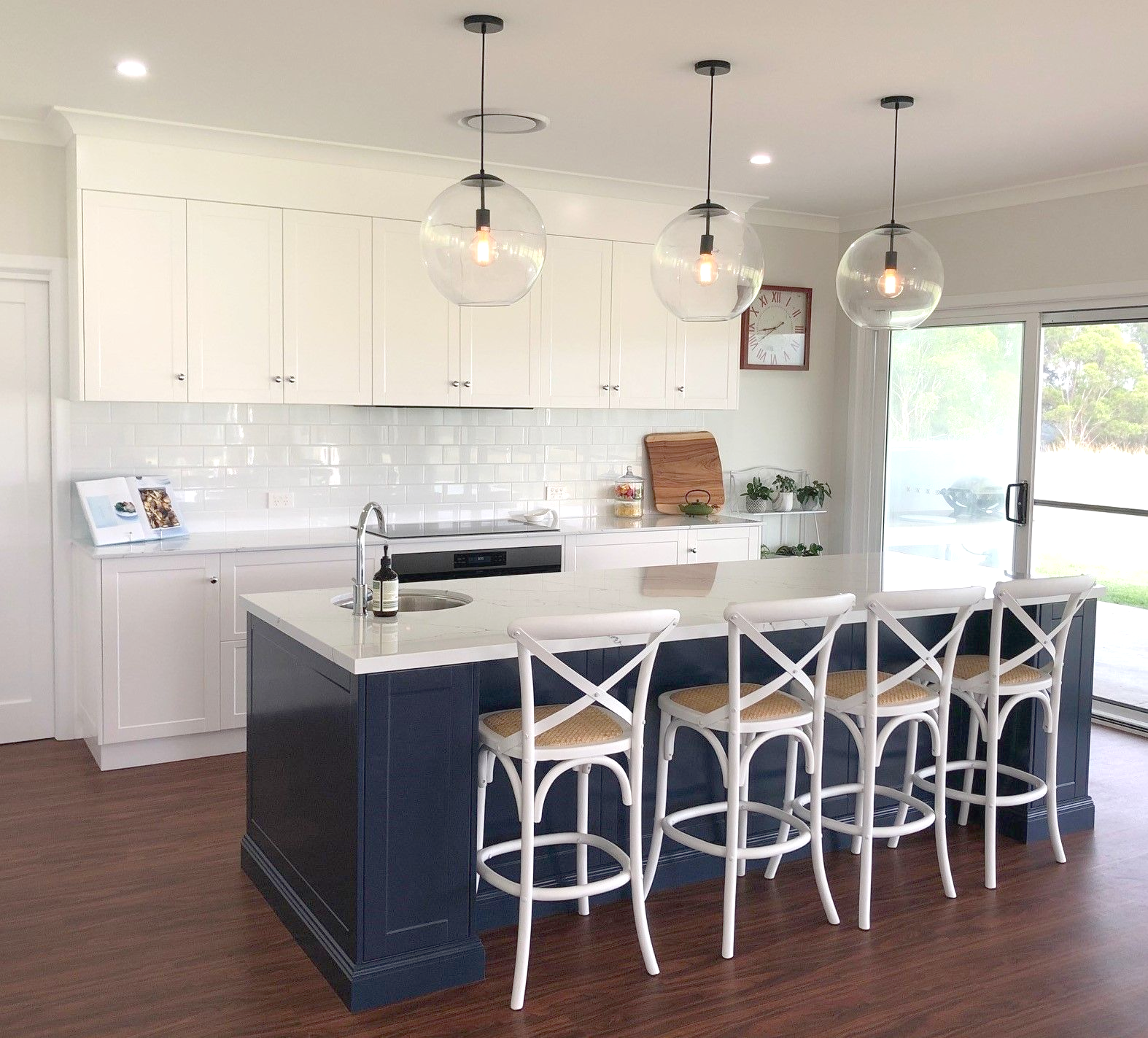 Modern kitchen with navy island, white cabinets, and clear globe pendant lights. White cross-back stools — Crispin Cabinetmakers in Grafton, NSW