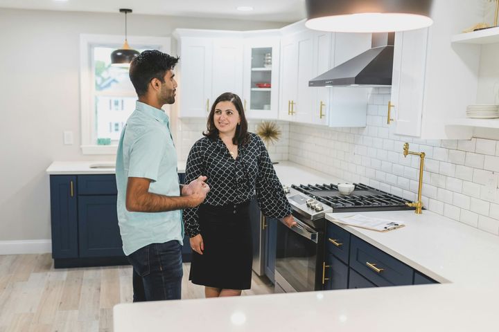 Man and woman in a renovated kitchen. The woman gestures near a stove, and they both appear to be in conversation.