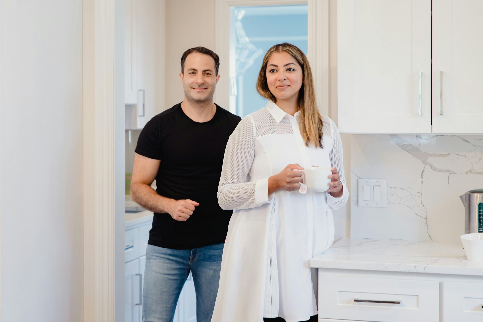 Man and woman in a kitchen, smiling. Woman holds a mug, wearing white. Man in black shirt and jeans. White cabinetry.