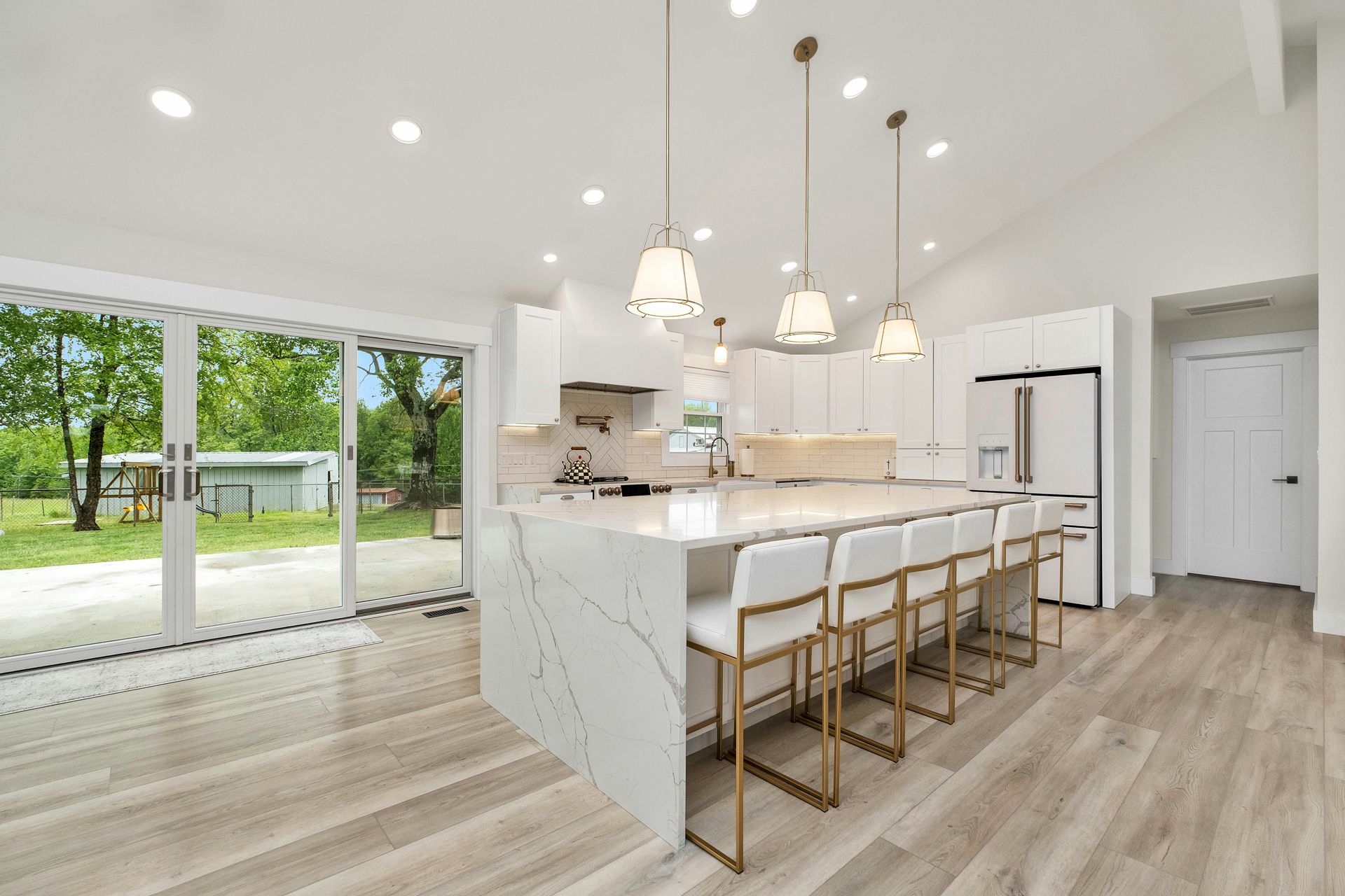 Modern white kitchen with island seating, pendant lights, and large sliding doors to a backyard.