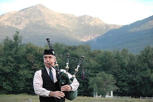 A man is playing a bagpipe in a field with mountains in the background.