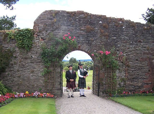 Two men playing bagpipes in front of a stone wall