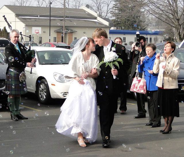 A bride and groom are walking in a parking lot while a man plays bagpipes