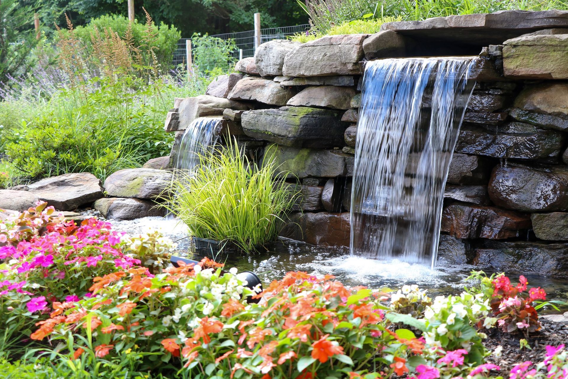 There is a waterfall in the middle of a garden surrounded by flowers.