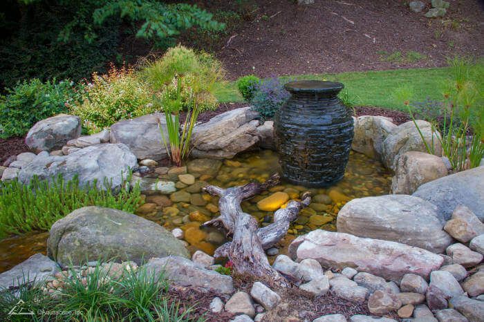 A pond surrounded by rocks and plants with a vase in the middle.