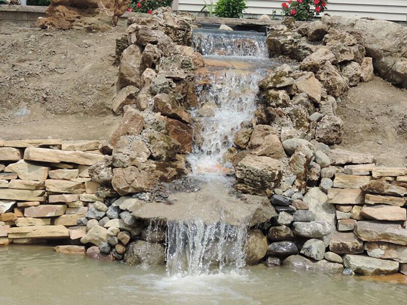 A small waterfall is surrounded by rocks and a brick wall.