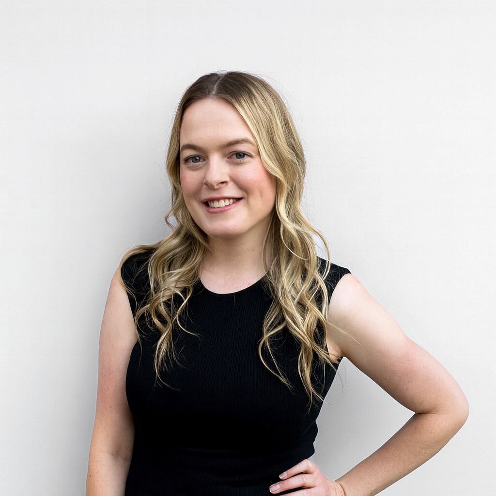 Woman with blonde hair smiling, wearing a black sleeveless top, hand on hip, leaning against a beige wall.