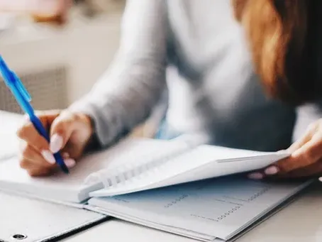 A woman is sitting at a table writing in a notebook with a pen.