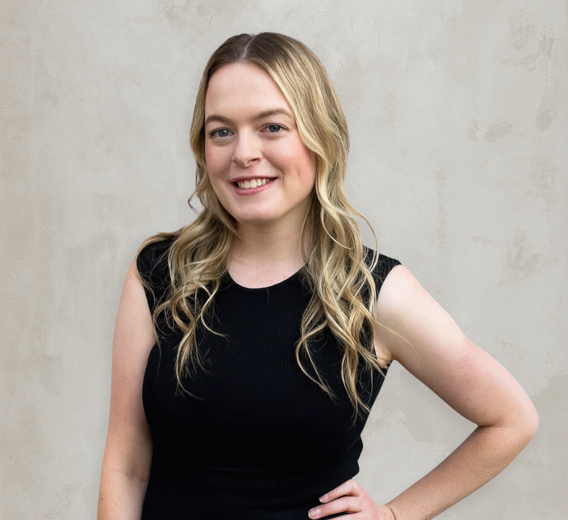 Woman with blonde hair smiling, wearing a black sleeveless top, hand on hip, leaning against a beige wall.