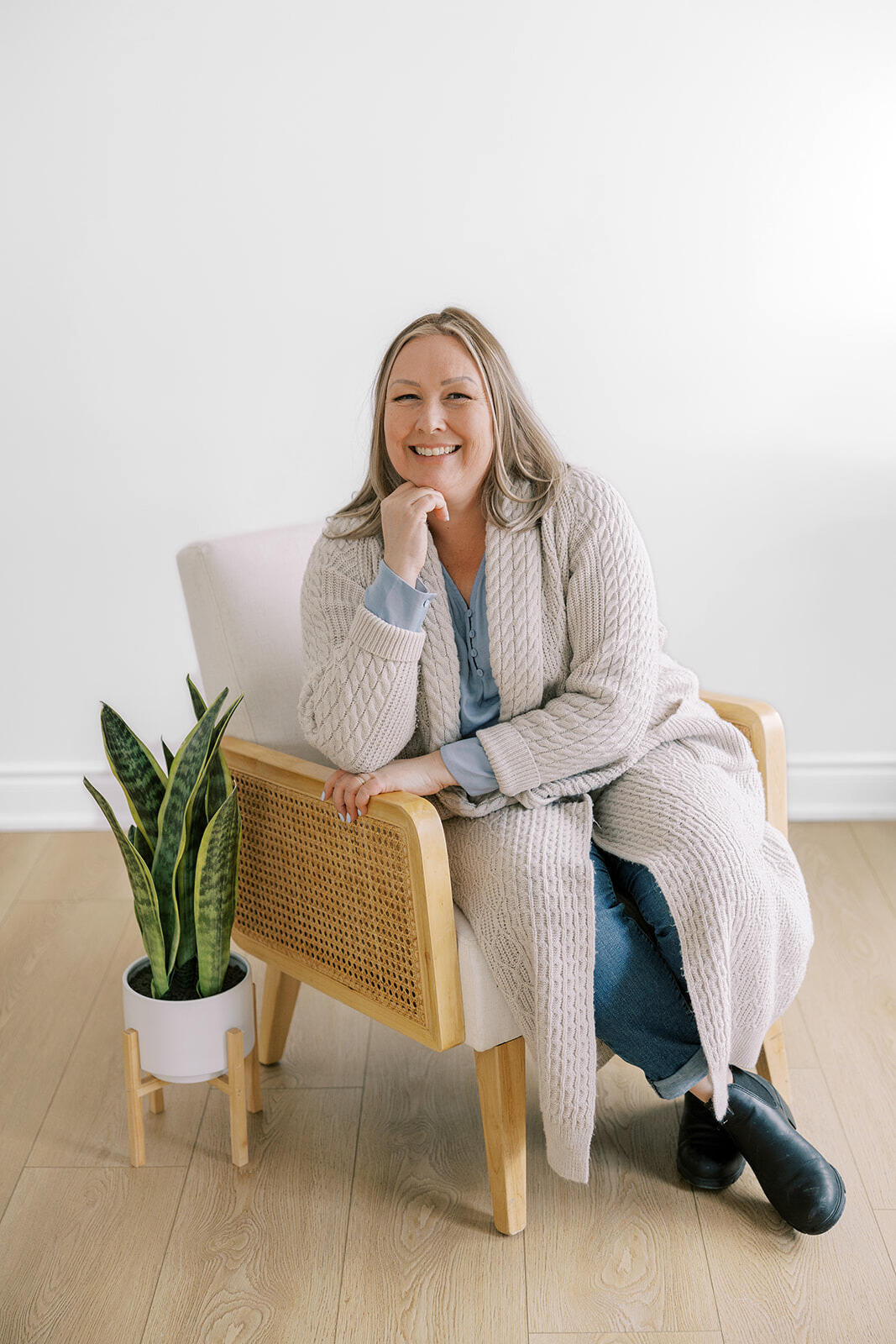 Woman in long cardigan sits in chair, smiling. Beside her is a snake plant in a white pot, on a wooden floor.