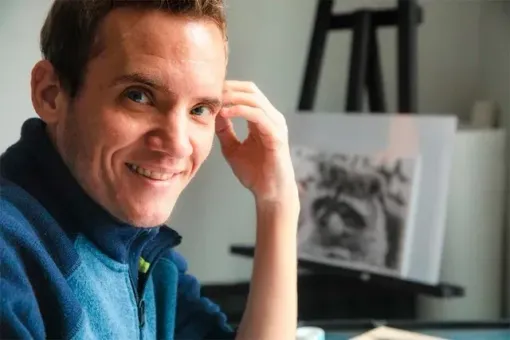 A man in a blue sweater is smiling while sitting at a table in front of an easel.
