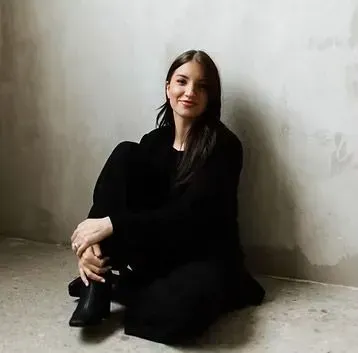 A woman in a black dress is sitting on the floor in front of a wall.