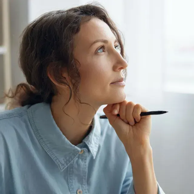 A woman is holding a pen to her chin and looking out a window.
