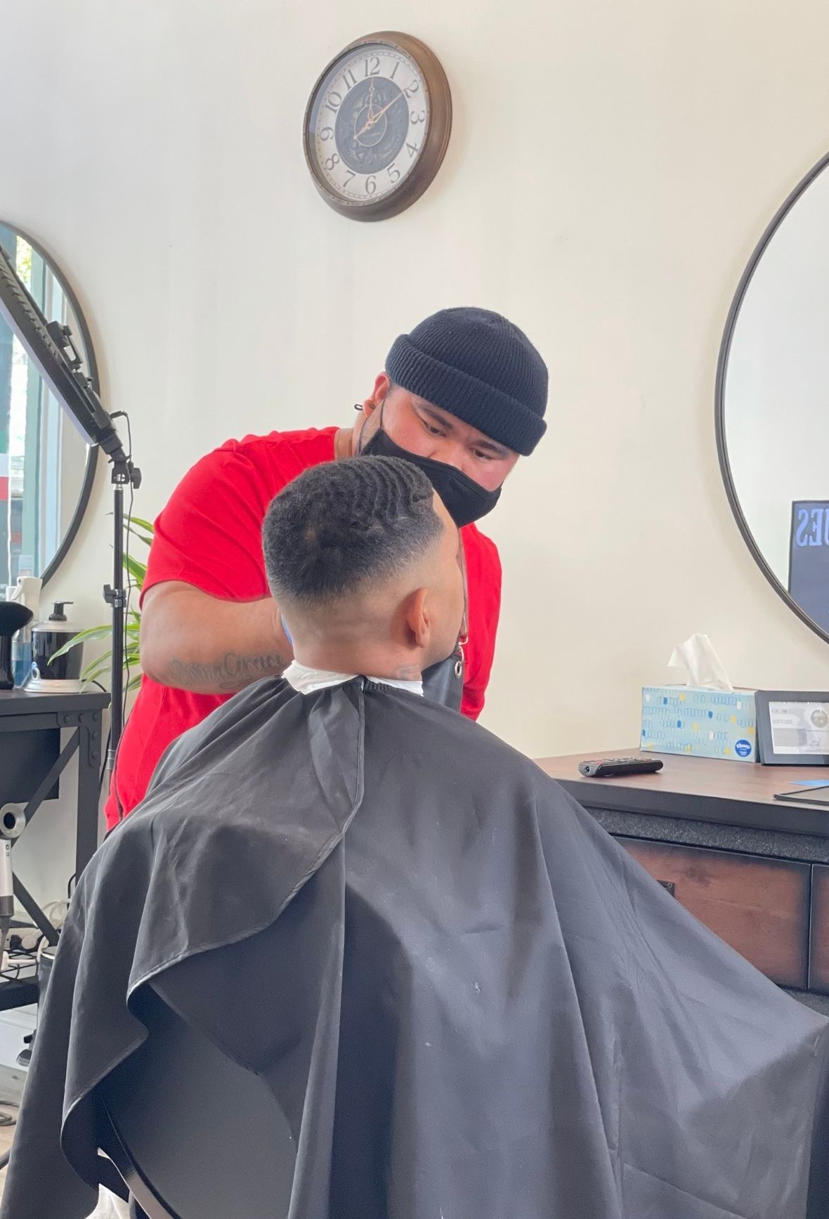 Barber giving a client a haircut in a shop. The barber wears a black cap and mask, the client a cape.