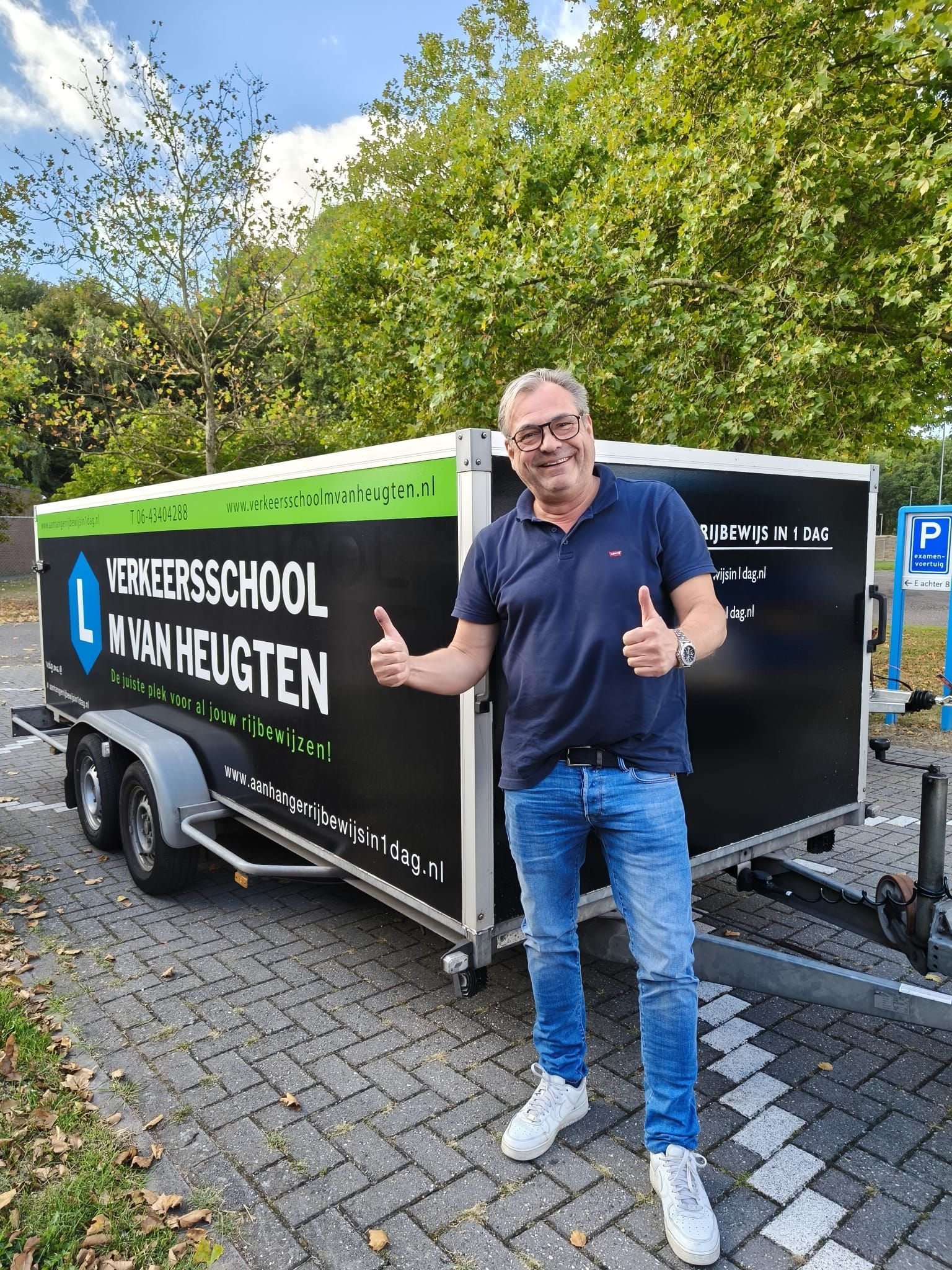 Een man steekt zijn duim omhoog naast een aanhanger met een zwart bord waarop staat: 