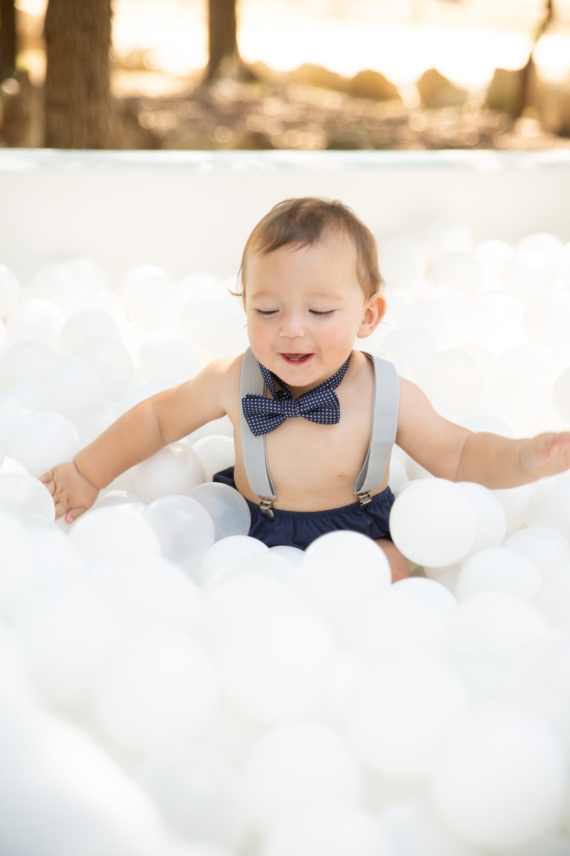 A baby wearing suspenders and a bow tie is sitting in a pool of white balloons.