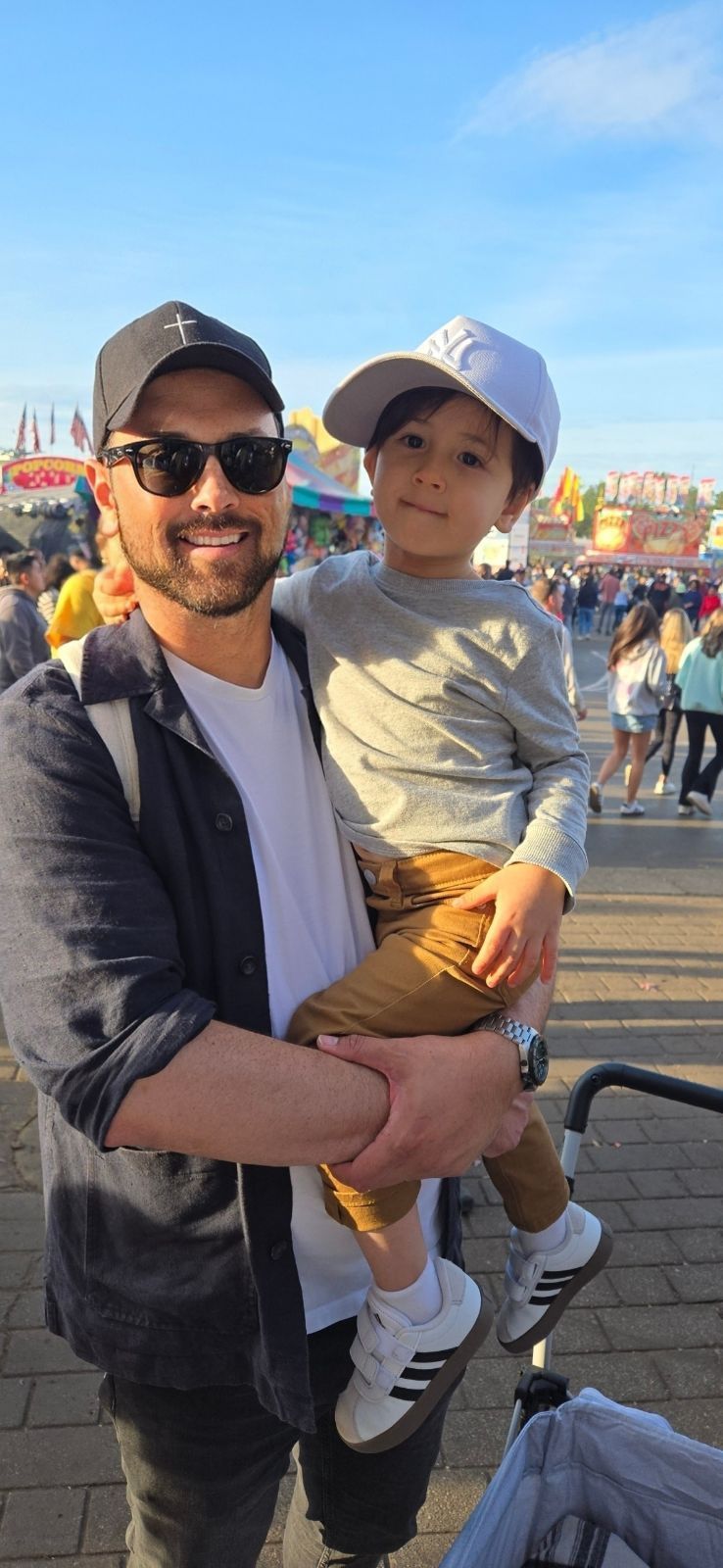 Del, in a dark cap and sunglasses, carries his kid ,in a white cap, outdoors.