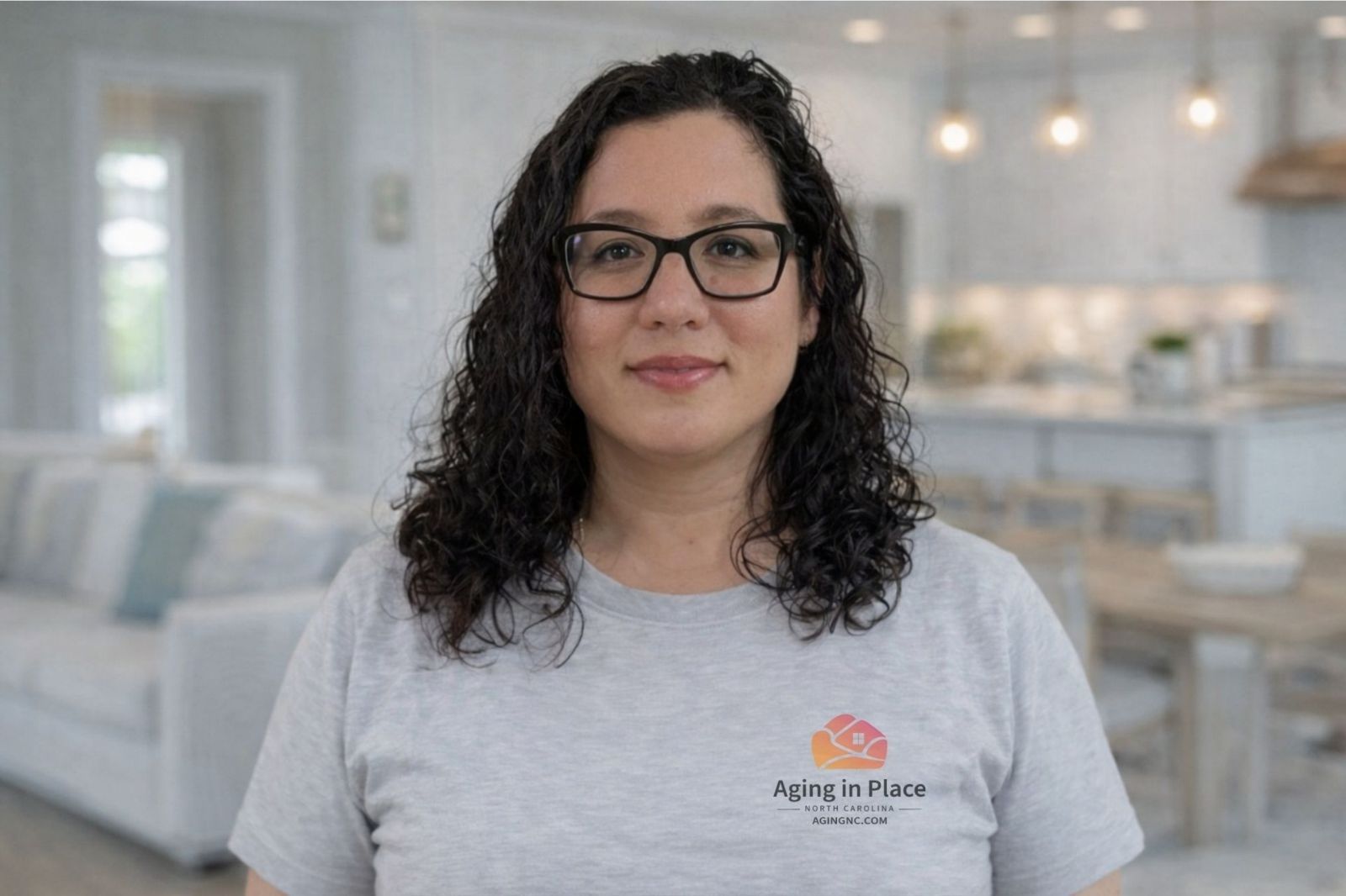 Mary in a gray t-shirt with an Aging in Place logo, standing in a brightly lit, blurred home interior.