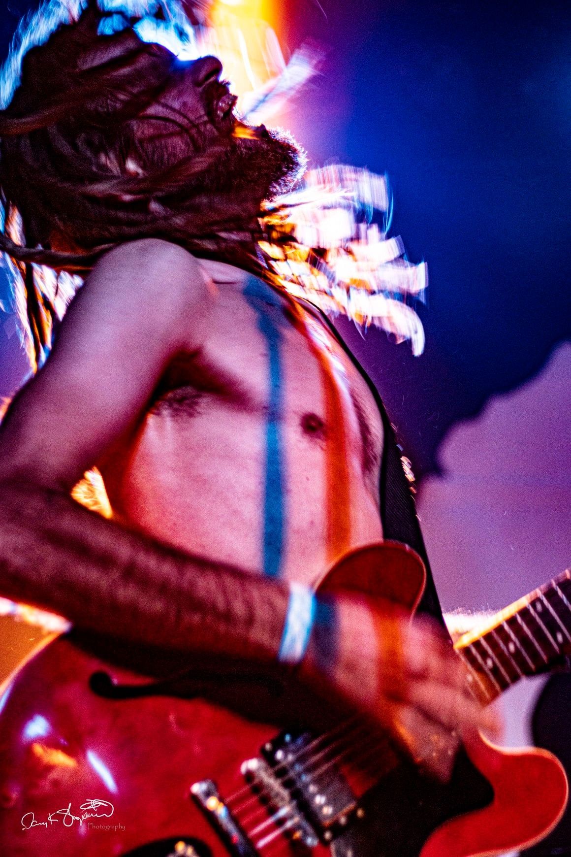 Shirtless guitarist with dreadlocks, playing a red electric guitar onstage, blue and red body paint, dynamic lighting.