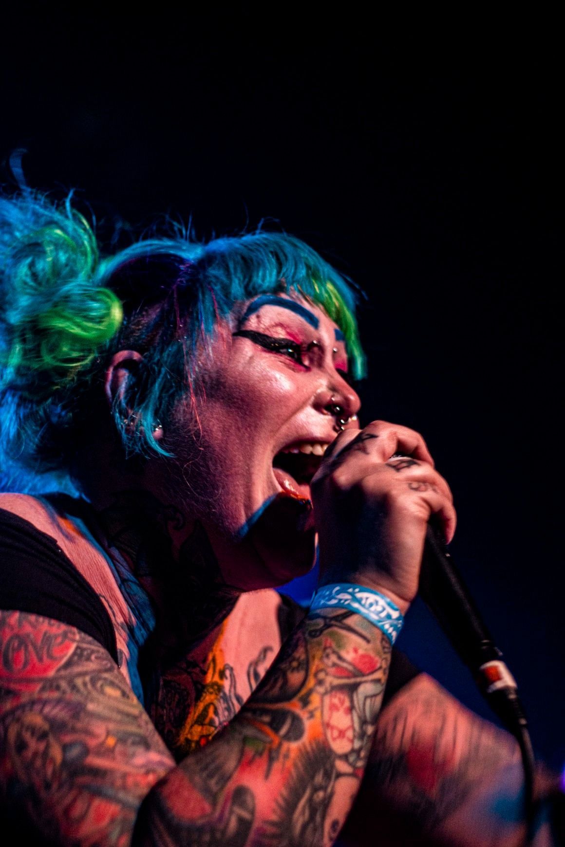 Person with blue and green hair, tattoos, and heavy makeup sings into a microphone with open mouth.