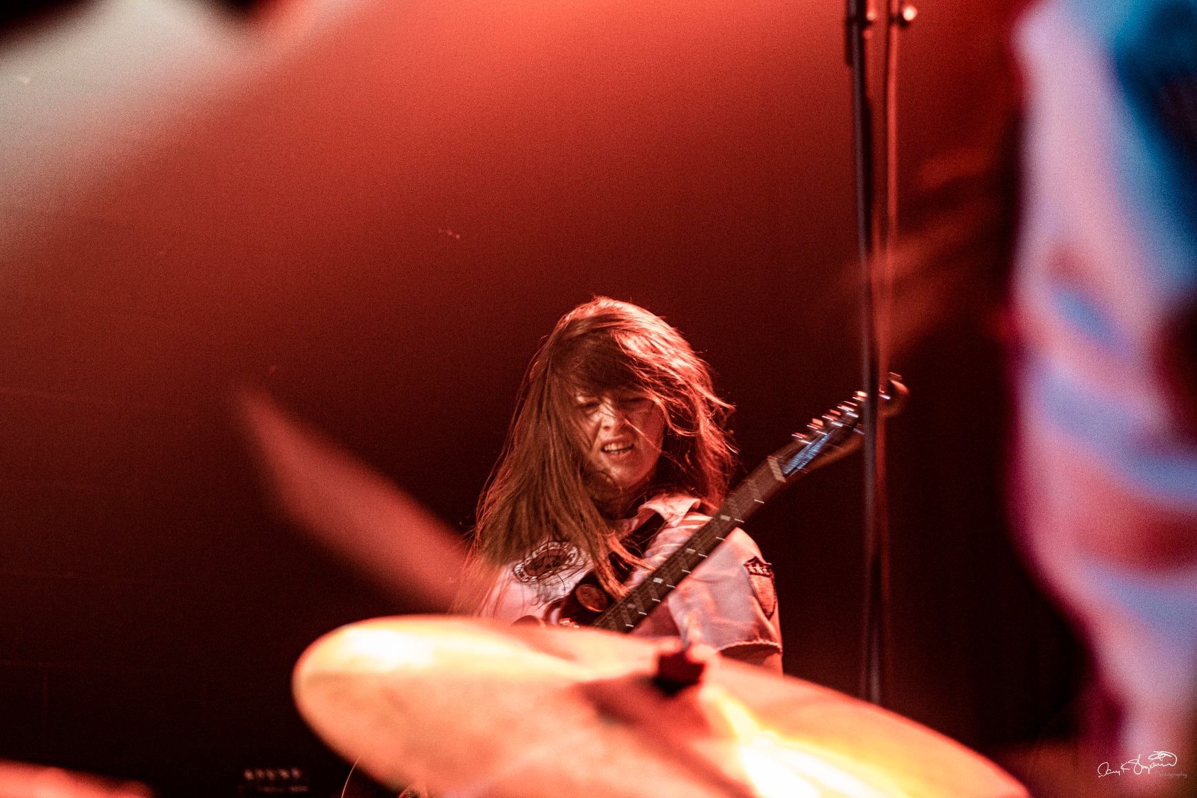 Guitarist on stage, lit by red light. Long hair, playing a guitar, with a drum cymbal in the foreground.