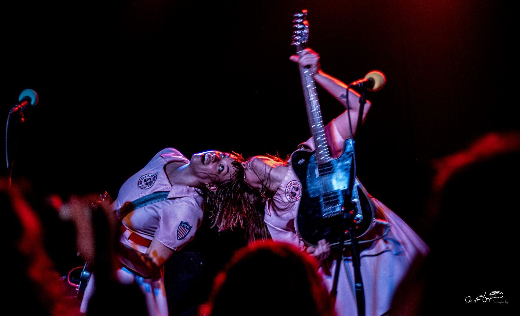 Woman playing guitar onstage, head thrown back, with microphone. Stage lit in red and blue.