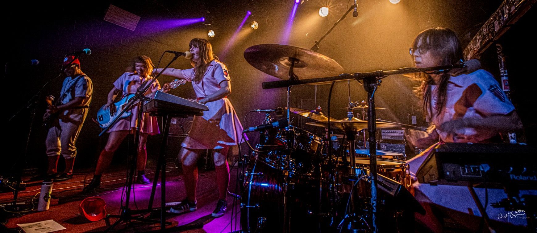 A band performs on stage. Musicians in light-colored outfits play instruments under stage lights.