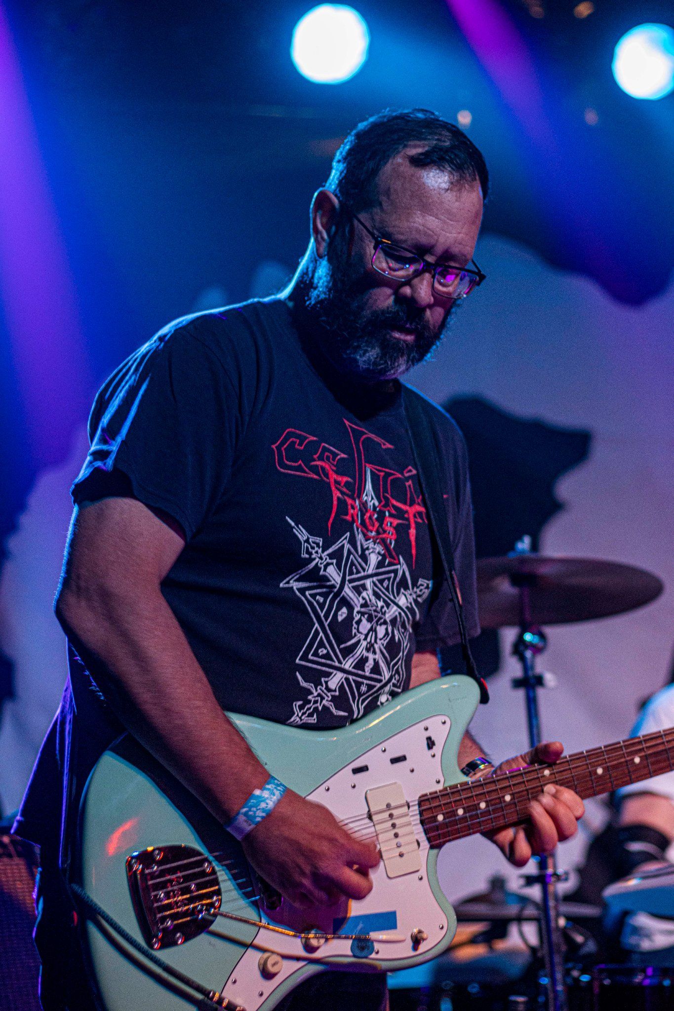 Man playing a light blue electric guitar on stage, wearing glasses and a t-shirt.