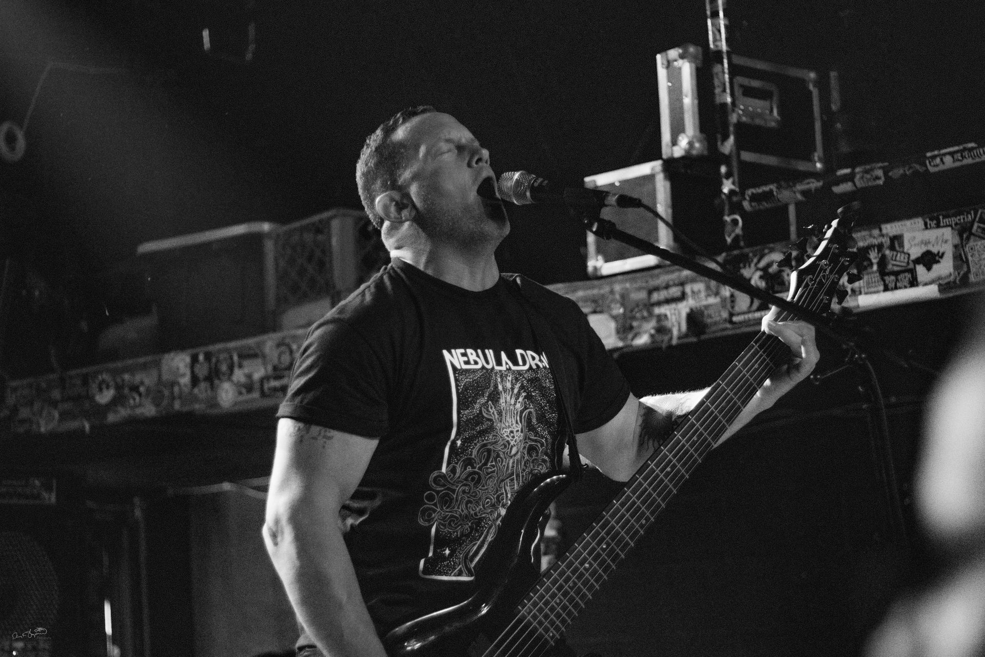 A musician playing bass and singing into a microphone on stage, black and white.