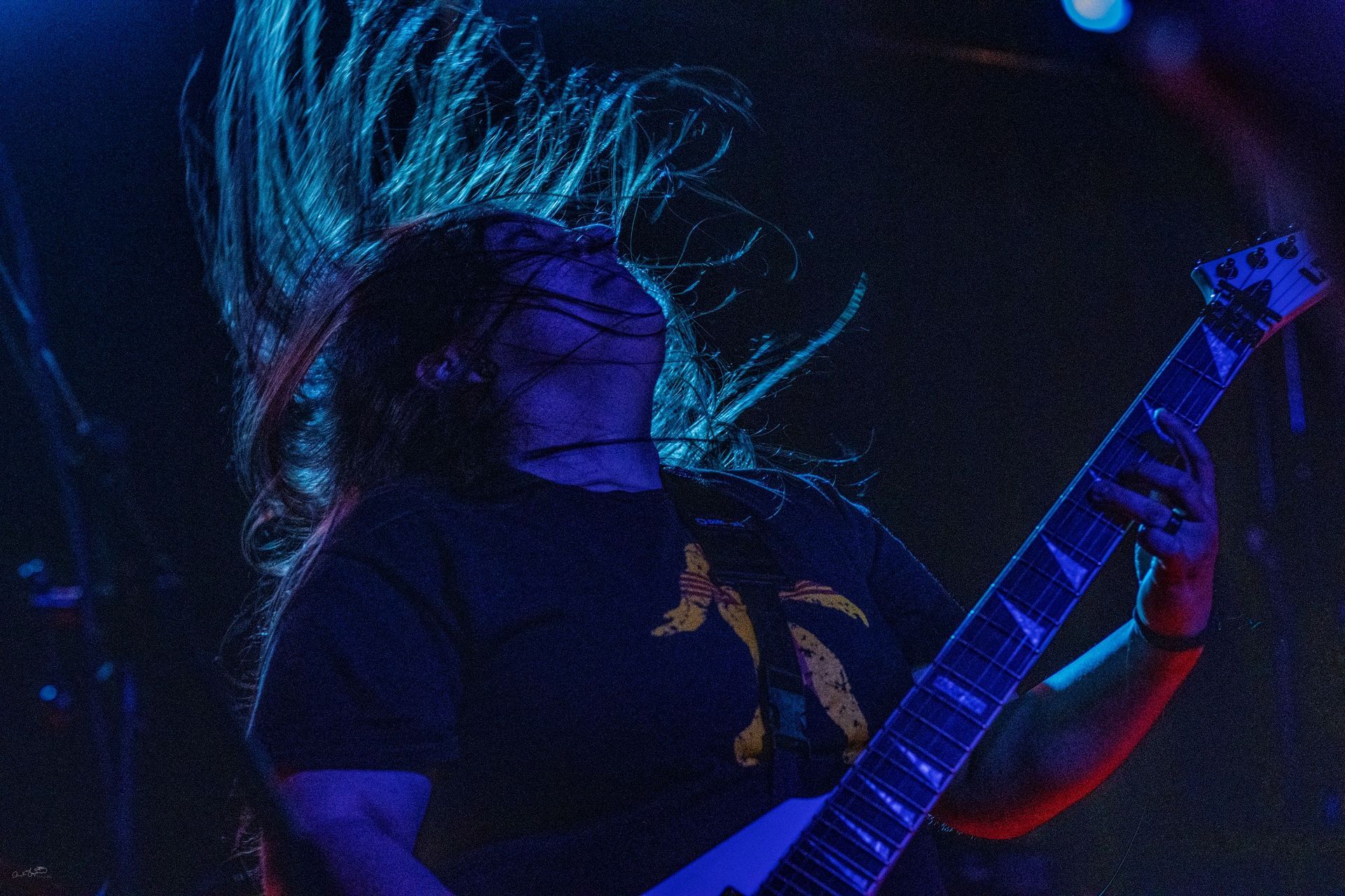 Guitarist with long hair, headbanging, playing a white V-shaped guitar on a dark stage.
