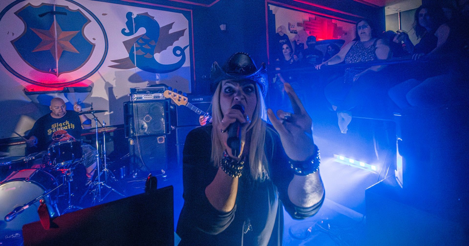 A musician with a small hat sings into a mic, making a rock hand gesture. A band plays in a dimly lit bar.